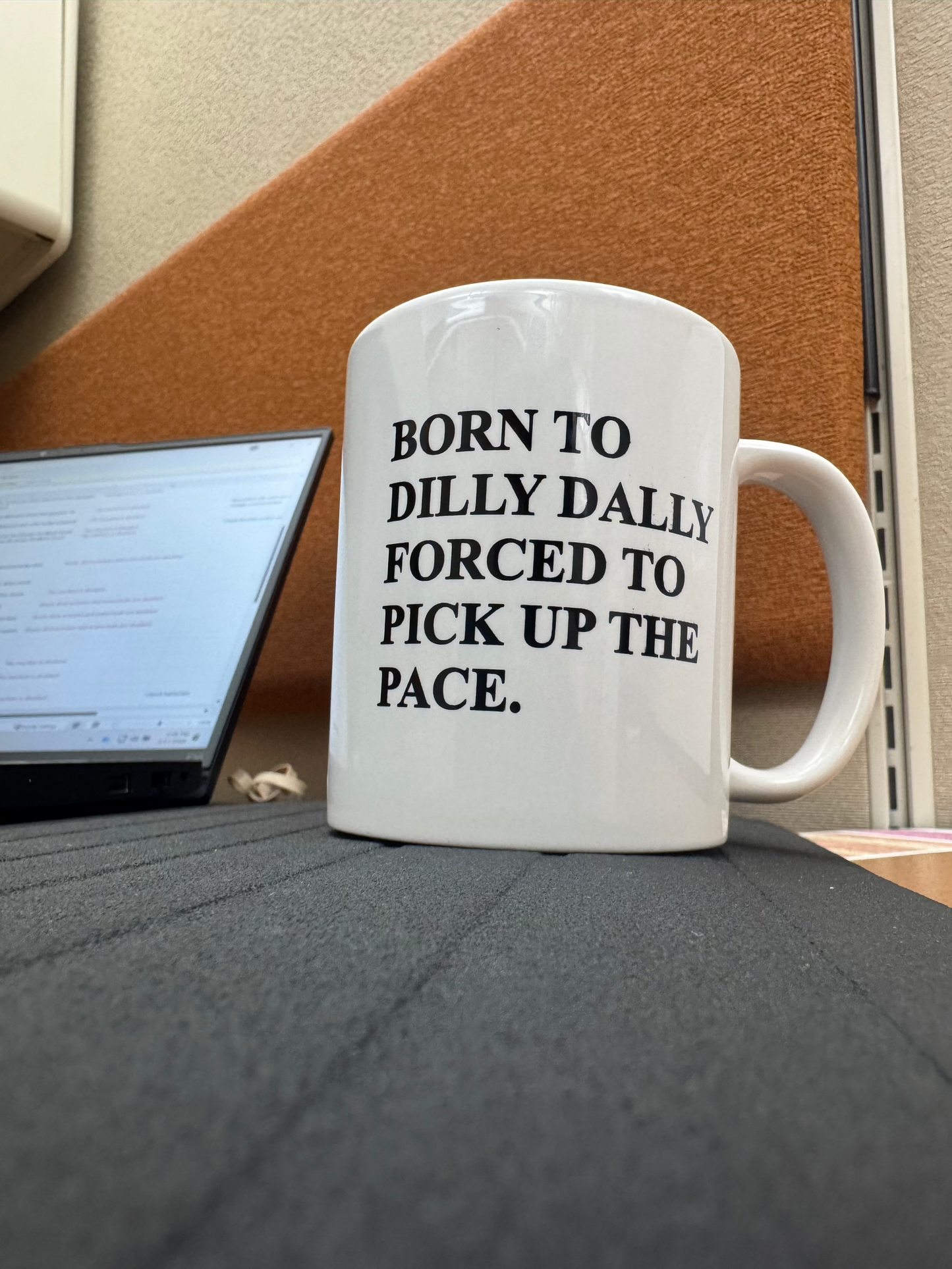 White mug with humorous text on a desk next to a laptop